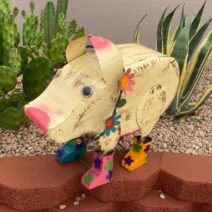 May include: A whimsical metal pig sculpture with a distressed white paint finish. The pig is wearing colorful boots and has floral accents.