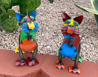 May include: Two colorful metal cat sculptures with whimsical details. The cats are painted in shades of green, blue, red, orange, and yellow. They have long, thin legs and a playful expression.