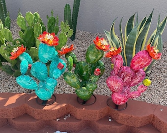 May include: Three metal cactus sculptures with colorful flowers. The cactus on the left is turquoise, the middle cactus is green, and the cactus on the right is pink.