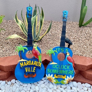 May include: Two blue painted metal guitars with colorful details. One guitar says "Margarita Ville" and the other says "It's 5 O'Clock Somewhere".