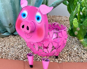 May include: Pink metal pig planter with a flower design. The pig has blue eyes and a smiling mouth.