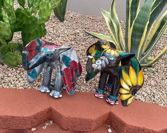 May include: Two colorful metal elephant sculptures with painted floral designs. The elephant on the left has red and blue flowers, while the elephant on the right has yellow sunflowers.