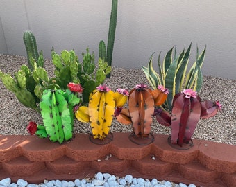 May include: Four metal cactus sculptures in different colors, green, yellow, orange, and red, with pink flowers. The sculptures are arranged in a row on a brown brick surface.