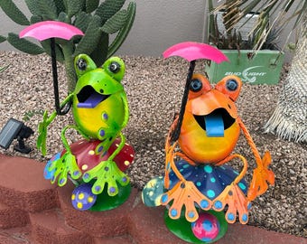 Può includere: Due figurine di rana in metallo colorate che tengono degli ombrelli. Le rane sono sedute su basi a forma di fungo con pois. Le rane sono verdi e arancioni con lingue blu.