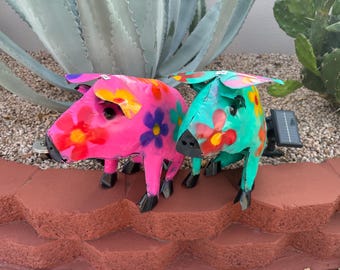 May include: Two metal pig figurines with colorful floral designs. The pig on the left is pink with orange, yellow, and blue flowers. The pig on the right is teal with red, yellow, and orange flowers.