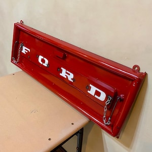 May include: Red metal tailgate with the word "FORD" in white letters. The tailgate has two metal hooks and a chain attached to the bottom.