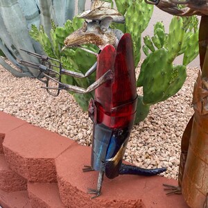 K&ouml;nnte beinhalten: Eine Metallskulptur einer Ratte, die einen Cowboyhut tr&auml;gt und eine Pistole h&auml;lt. Die Ratte ist rot, blau und goldfarben bemalt. Die Skulptur steht auf einer roten Backsteinmauer.