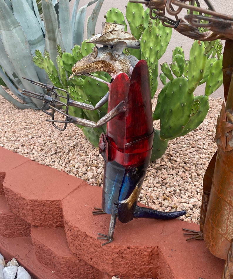 K&ouml;nnte beinhalten: Eine Metallskulptur einer Ratte, die einen Cowboyhut tr&auml;gt und eine Pistole h&auml;lt. Die Ratte ist rot, blau und goldfarben bemalt. Die Skulptur steht auf einer roten Backsteinmauer.