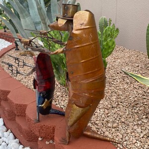 K&ouml;nnte beinhalten: Zwei Armadillo-Skulpturen aus Metall, eine mit Hut, die eine Waffe h&auml;lt, und die andere in Rot, Wei&szlig; und Blau. Die Skulpturen stehen auf einer roten Ziegelstein-Oberfl&auml;che.