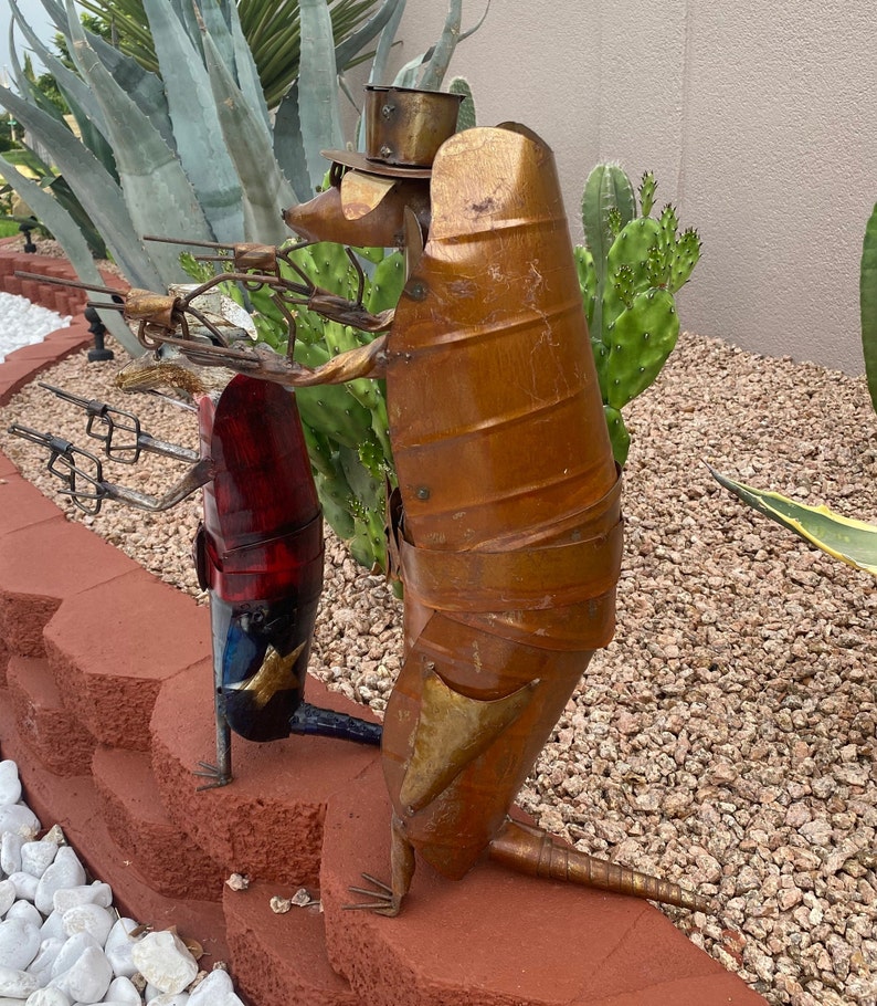 K&ouml;nnte beinhalten: Zwei Armadillo-Skulpturen aus Metall, eine mit Hut, die eine Waffe h&auml;lt, und die andere in Rot, Wei&szlig; und Blau. Die Skulpturen stehen auf einer roten Ziegelstein-Oberfl&auml;che.