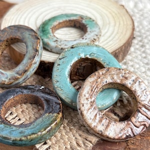 May include: A collection of handmade ceramic beads in various shapes and colors. The beads are glazed in shades of blue, green, and brown, with a rustic, textured finish. Some beads are oval, while others are circular, with a hole in the center.
