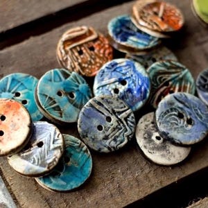 May include: A collection of handmade ceramic buttons in various colors, including blue, orange, and white. Each button has two holes and unique patterns, resting on a rustic wooden surface. These decorative buttons are ideal for sewing and craft projects.