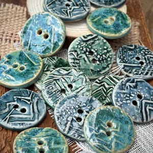 May include: A collection of round ceramic buttons featuring two holes in the center. Each button displays a unique glaze pattern in shades of teal, blue, and green, with some showing textured, raised lines and others featuring speckled, mottled finishes.