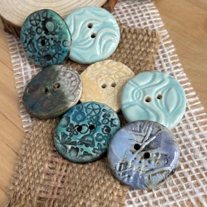 May include: A collection of handmade ceramic buttons in various colors and designs. The buttons feature floral, swirl, and abstract patterns. Colors include teal, light blue, beige, and a marbled blue-purple. They are arranged on a burlap and white fabric background.