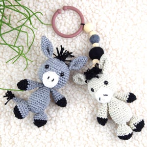 Könnte beinhalten: Ein graues gehäkeltes Eselspielzeug mit schwarzen Hufen und einer schwarzen Mähne. Der Esel ist an einem Mobile aus Holzperlen und grauen Perlen mit einem rosa Plastikring befestigt.