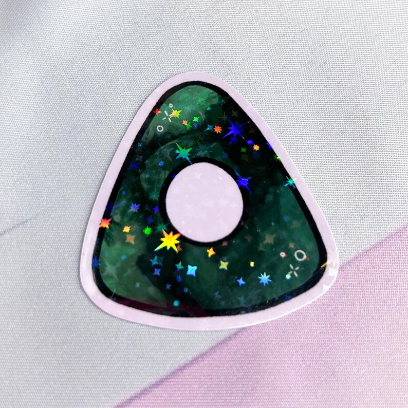 K&ouml;nnte beinhalten: Ein gr&uuml;ner holografischer Ouija-Planchette-Aufkleber mit einem wei&szlig;en Kreis in der Mitte und einem schwarzen Umriss.