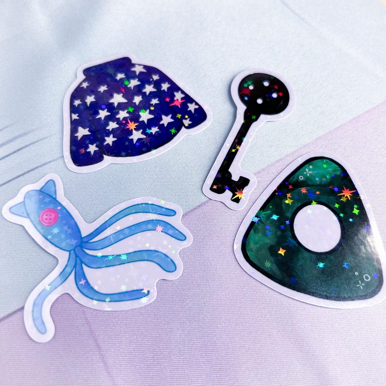 K&ouml;nnte beinhalten: Vier holografische Aufkleber mit einem blauen Pullover mit wei&szlig;en Sternen, einem schwarzen Schl&uuml;ssel, einem blauen Tintenfisch und einem gr&uuml;nen Geist mit einem wei&szlig;en Kreis in der Mitte.