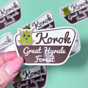 May include: A collection of stickers featuring a brown and white sign design. The sign reads "Korok Great Hyrule Forest" in a stylized font, with a green leaf character illustration. The stickers are on a light green background.