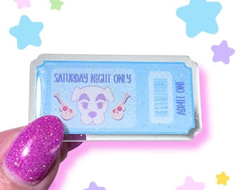 May include: A light blue ticket-shaped pin with glitter accents. The pin features a cartoon dog, two guitars, and the text "SATURDAY NIGHT ONLY" and "ADMIT ONE". The pin is held by a hand with a sparkly pink nail.