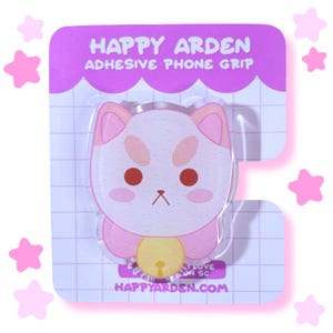 May include: A Happy Arden adhesive phone grip featuring a glittery, light pink cat character with dark red eyes and a yellow bell. The item is mounted on a purple and white grid-patterned backing card with the text Happy Arden Adhesive Phone Grip.