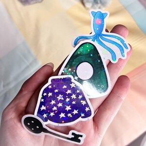K&ouml;nnte beinhalten: Ein Set aus vier holografischen Aufklebern mit einem gr&uuml;nen und wei&szlig;en Dreieck, einem blauen und wei&szlig;en Oktopus, einem lila und wei&szlig;en sternenf&ouml;rmigen Umhang und einem schwarzen und wei&szlig;en Schl&uuml;ssel.