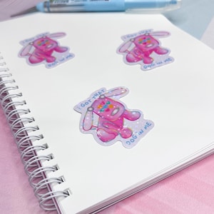 May include: Three holographic stickers featuring a pink cartoon dog with the text "I GOT THAT DOG IN ME". The stickers are on a white spiral-bound notebook with a pink background and a blue pen.