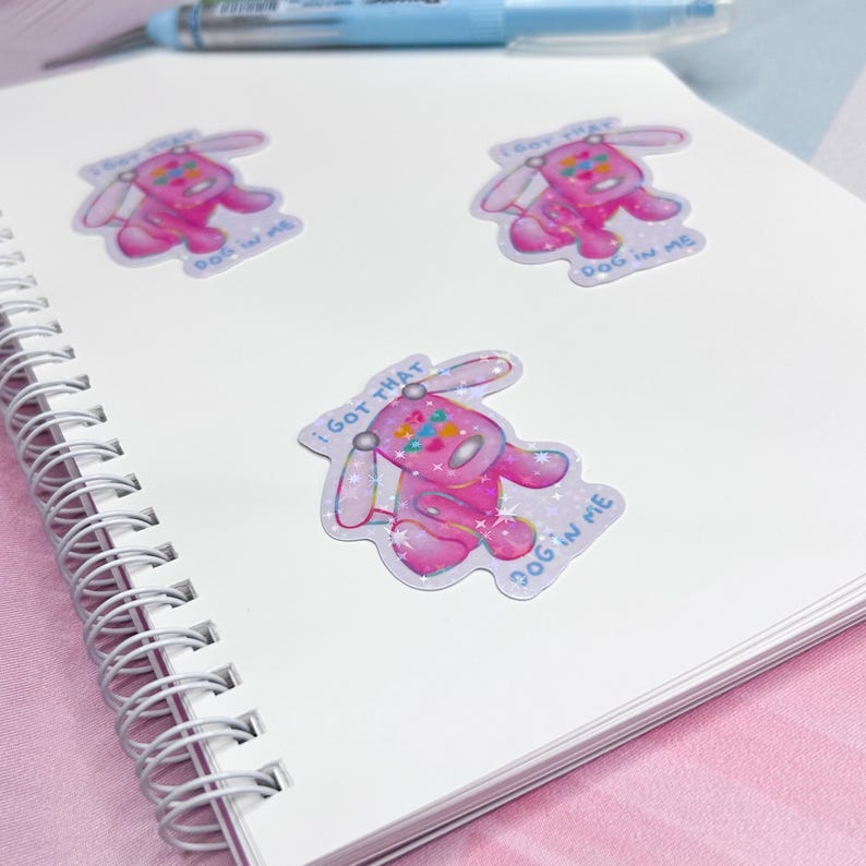 May include: Three holographic stickers featuring a pink cartoon dog with the text "I GOT THAT DOG IN ME". The stickers are on a white spiral-bound notebook with a pink background and a blue pen.