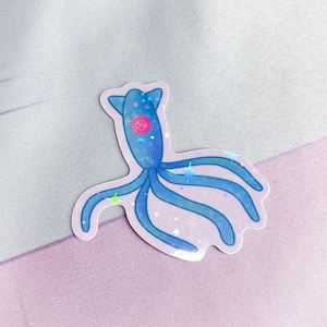 K&ouml;nnte beinhalten: Ein blauer, schimmernder Aufkleber mit einer Cartoon-Oktopus mit einem rosa Knopf als Auge. Der Oktopus hat sechs Tentakel und ist von schimmernden, regenbogenfarbenen Sternen umgeben.