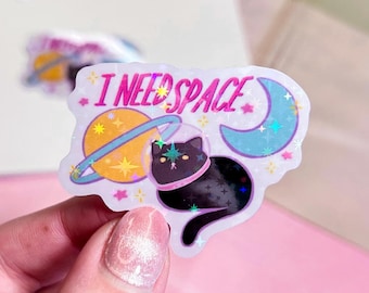May include: A holographic sticker with a black cat wearing a space helmet, a planet, a crescent moon, and the text "I NEED SPACE".