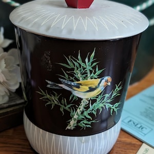 May include: A cylindrical metal canister with a black body and a light gray base and lid. The lid features a red hexagonal knob and a white zigzag pattern. The front displays a colorful illustration of a goldfinch perched on a green branch.