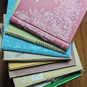 May include: A stack of vintage books with colorful covers. The top book is a dusty rose color with white floral illustrations and the title "Wild Flowers of the World." Other books include titles like "EHRMANN" and "HUMMEL."