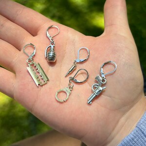 May include: Five silver dangle earrings with different charms. The charms include a razor blade, a grenade, a bullet, handcuffs, and a pistol.