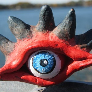 Op de afbeelding: Een handgemaakte, grillige sculptuur van een oog. Het oog is wit met een blauwe iris en een zwarte pupil. Het is geplaatst in een rode, open mondvorm met zwarte, vingerachtige uitsteeksels.