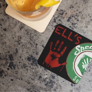 May include: Black square coaster featuring a red bloody handprint and a green logo that reads "Ell's Special Sauce".