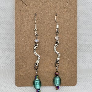 May include: A pair of handcrafted earrings with a silver-tone metal design. Each earring features a wavy, textured silver element, clear and iridescent beads, and a teal-colored glass bead with purple accents. The earrings hang from silver-tone ear wires.