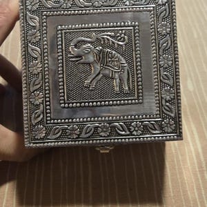 May include: A square silver-colored metal box featuring an embossed elephant design in the center. The lid is decorated with a detailed border of floral patterns and leaves, surrounded by a beaded frame that highlights the central elephant motif.