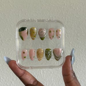 May include: A clear plastic case with 10 false nails. The nails are decorated with green, gold, and silver designs. Some nails have a swirl design, others have stars, and some have a green and silver design.
