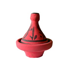 Op de afbeelding: Een rode keramische tajinepot met een conisch deksel en een ronde basis. De pot heeft zwart geschilderde decoratieve patronen rond het deksel. De tajine is een traditioneel kookvat dat in de Noord-Afrikaanse keuken wordt gebruikt.