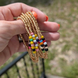 May include: Gold beaded bracelets with colourful accent beads. The bracelets are held in a hand with orange nail polish. The accent beads are pink, yellow, green, purple, red, black, and white.
