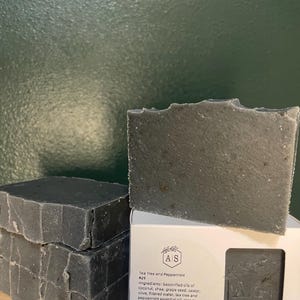 May include: Several dark gray soap bars are stacked and displayed next to a white box. The box has the text "Tea Tree and Peppermint #29" and lists ingredients. The soap bars are approximately 5 oz each.