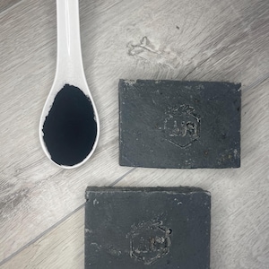 Può includere: Due saponette rettangolari al carbone grigio scuro, ciascuna con un logo esagonale stampato con le lettere AIS, posizionate accanto a un cucchiaio di ceramica bianca contenente un mucchietto di polvere di carbone nera fine.