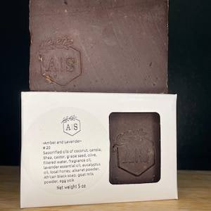 May include: A dark brown soap bar with a stamped design and a white box. The box has a label with the text "Amber and Lavender" and a list of ingredients. The soap weighs 5 oz.