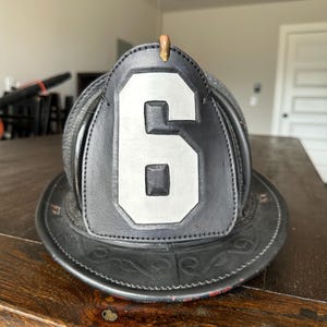 May include: A black leather firefighter helmet with a silver number 6 on the front. The helmet has a decorative brim and a brass fitting on top. The helmet is sitting on a wooden surface.