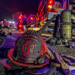 May include: A red and black firefighter helmet with the text "MAPLE STREET MAFIA" and "LUCIAN" is in the foreground. A fire truck and other emergency personnel are visible in the background, illuminated by red and blue lights. The scene is set at night.