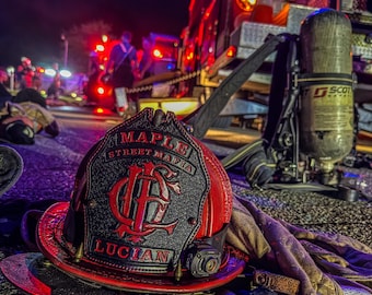 May include: A red and black firefighter helmet with the text "MAPLE STREET MAFIA" and "LUCIAN" is in the foreground. A fire truck and other emergency personnel are visible in the background, illuminated by red and blue lights. The scene is set at night.