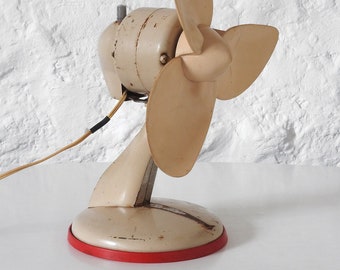 May include: A vintage, off-white desk fan with three blades. The fan has a red base and a cord.