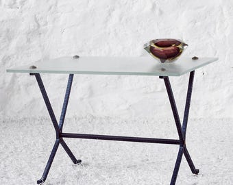 May include: A glass-topped table with a black metal frame. The table has a simple, modern design with a rectangular top and an X-shaped base. The table is sitting on a white surface.