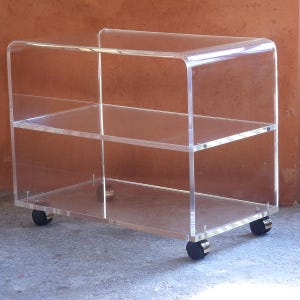以下が含まれることがあります： 3段の透明アクリル製ローリングカート。カートは湾曲したハンドルを持ち、4つの黒いキャスターホイールの上にあります。棚は透明で、ユニット全体を見ることができます。保管と展示用に設計されています。