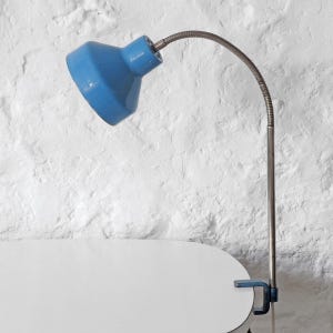 Può includere: Una lampada da scrivania blu vintage con un braccio a collo di cigno flessibile color argento. La lampada ha una base a morsetto ed è posizionata contro un muro bianco strutturato. Il paralume è blu brillante.
