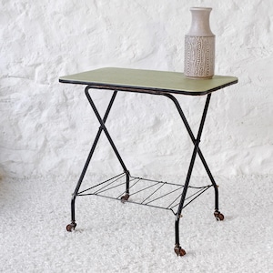 Może przedstawiać: Wózek do serwowania w stylu vintage z jasnozielonym prostokątnym blatem i czarną metalową ramą. Wózek ma dolną półkę i małe kółka. Na górze stoi beżowy wazon z teksturowanym wzorem.